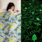 Girl sleeping under a light blue Famyo dinosaur-themed glow-in-the-dark blanket, shown glowing green with dinosaur and leaf patterns in the dark.
