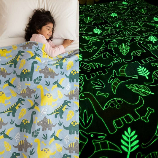 Girl sleeping under a light blue Famyo dinosaur-themed glow-in-the-dark blanket, shown glowing green with dinosaur and leaf patterns in the dark.