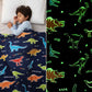 Child sleeping under a navy blue Famyo dinosaur-themed glow-in-the-dark blanket, shown glowing with green dinosaur outlines in the dark.