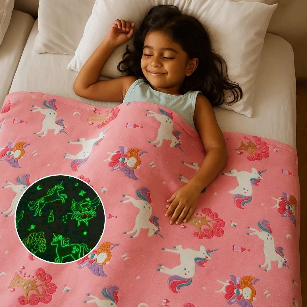 A young Indian girl is peacefully sleeping on a white bed, smiling softly while covered with a pink glow-in-the-dark unicorn blanket featuring fairies, castles, and crowns. An inset circle shows the blanket’s glowing design in the dark, revealing bright green outlines of unicorns and fairies.