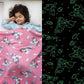 Girl sleeping under a pink Famyo glow-in-the-dark blanket with unicorn and cloud patterns, glowing green in the dark.